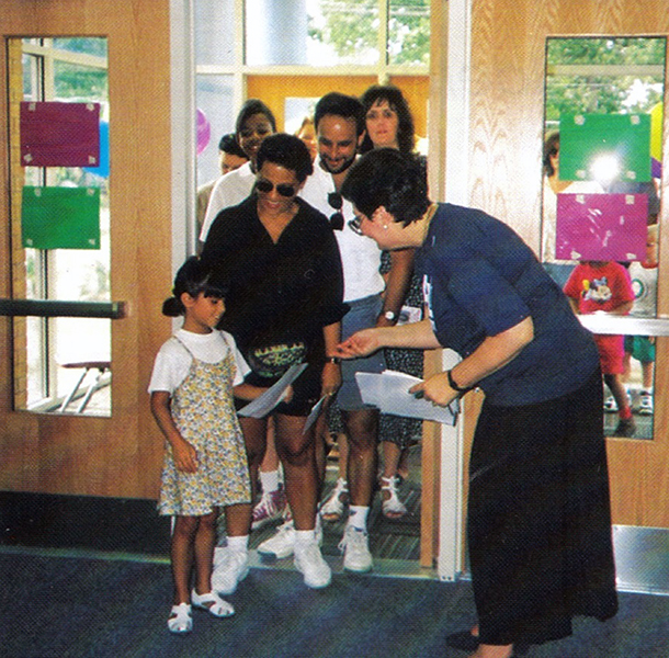 Color photograph taken from inside the main entrance at Lane Elementary. Principal Brower is standing in the doorway greeting a student. Her parents stand behind her, smiling broadly. More children and parents are visible in the background outside. Principal Brower holds a stack of papers in one hand, and has handed the girl a piece of paper.