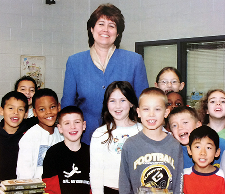 Color portrait of Principal Montgomery from our 2005 to 2006 yearbook. She is pictured standing behind a group of approximately 11 students. Everyone pictured is smiling broadly at the camera. 