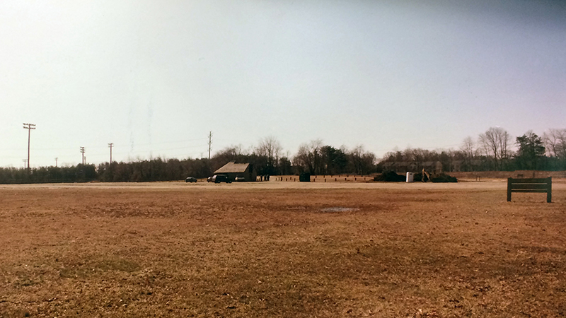 Exterior view of the future site of Lane Elementary School taken on March 11, 1994 prior to groundbreaking. The landscape is a grassy field. The grass is brown after having being dormant during the winter. A small building and sign are visible in the distance.  