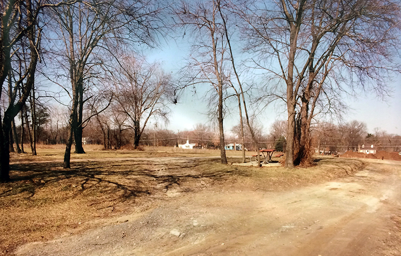 Exterior view of the future site of Lane Elementary School taken on March 11, 1994 prior to groundbreaking. The land is a park with small clusters of trees. There are dirt trails and a dirt driveway running through the property. A picnic table is set on a concrete pad amidst a small cluster of trees at center. A church and other buildings are pictured in the far distance on the opposite side of Beulah Street. The trees have all shed their leaves and lie dormant after the winter.  