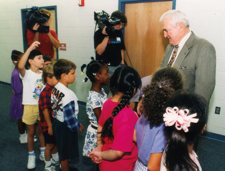 Color photograph of Anthony Lane standing in a hallway at Lane Elementary on opening day. He is speaking with a group of nine students. FCPS Red Apple 21 news crew cameramen are in the background. 