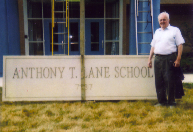 The stone entrance sign is positioned on the ground in front of the school’s main entrance. Anthony Lane is standing next to it, smiling for the camera. Ladders can be seen in the background, and a heavy duty strap or rope is attached around the center of the sign, ready to hoist it into the air. 